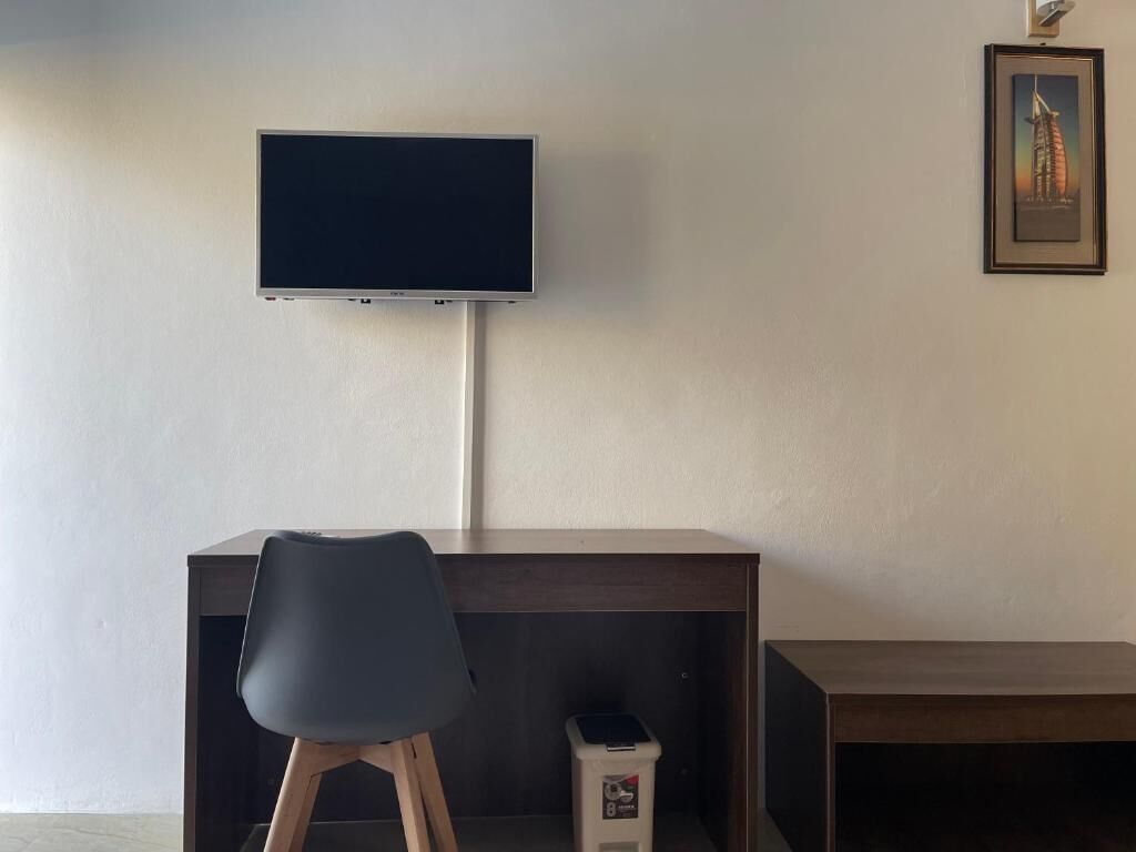 A room with a desk, a chair, a television mounted on the wall, and a trash bin.