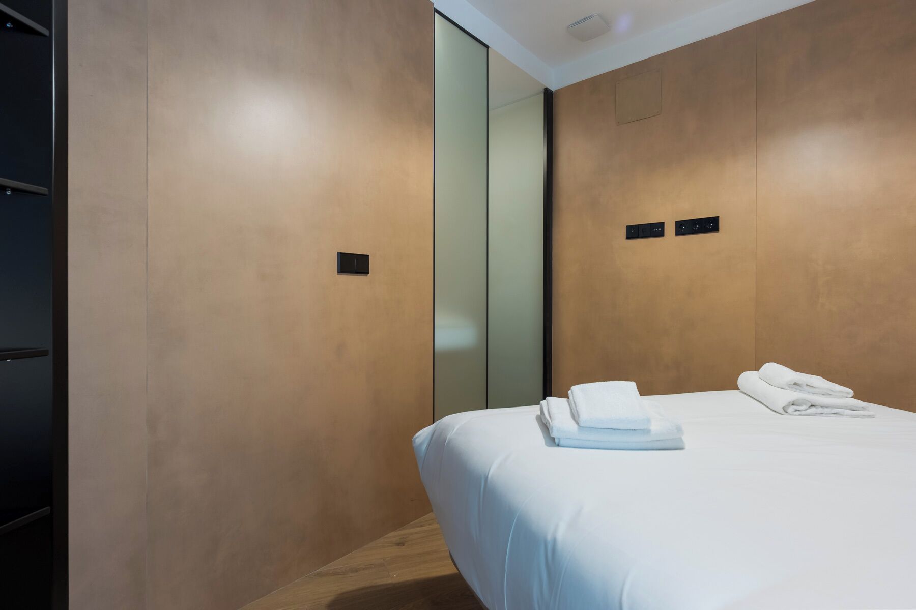 A modern hotel room with a bed, a small bedside table, and a wall-mounted control panel.