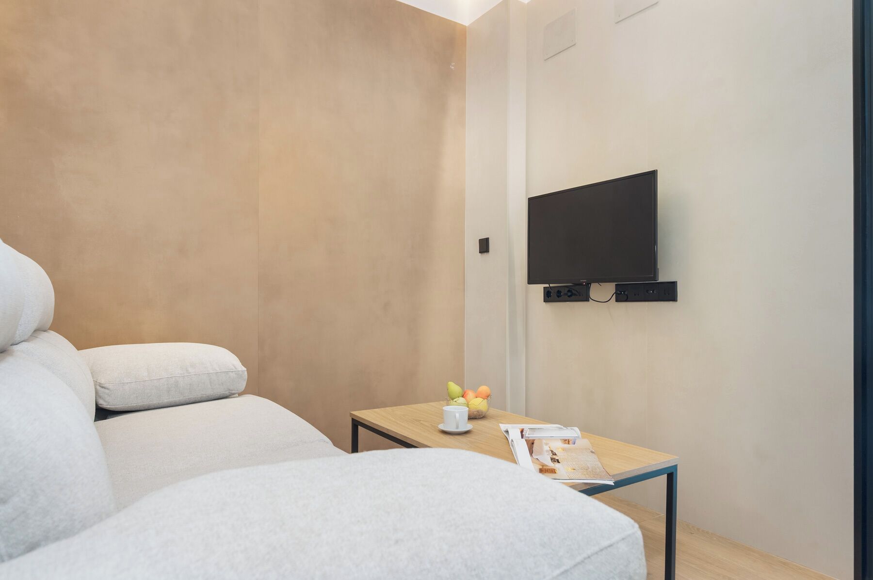 Een moderne kamer met een bank, een klein tafeltje met een kopje en fruit, en een aan de muur bevestigde televisie.