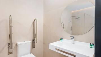 Un baño moderno con inodoro, ducha con barra de apoyo, lavamanos con espejo y artículos de aseo.