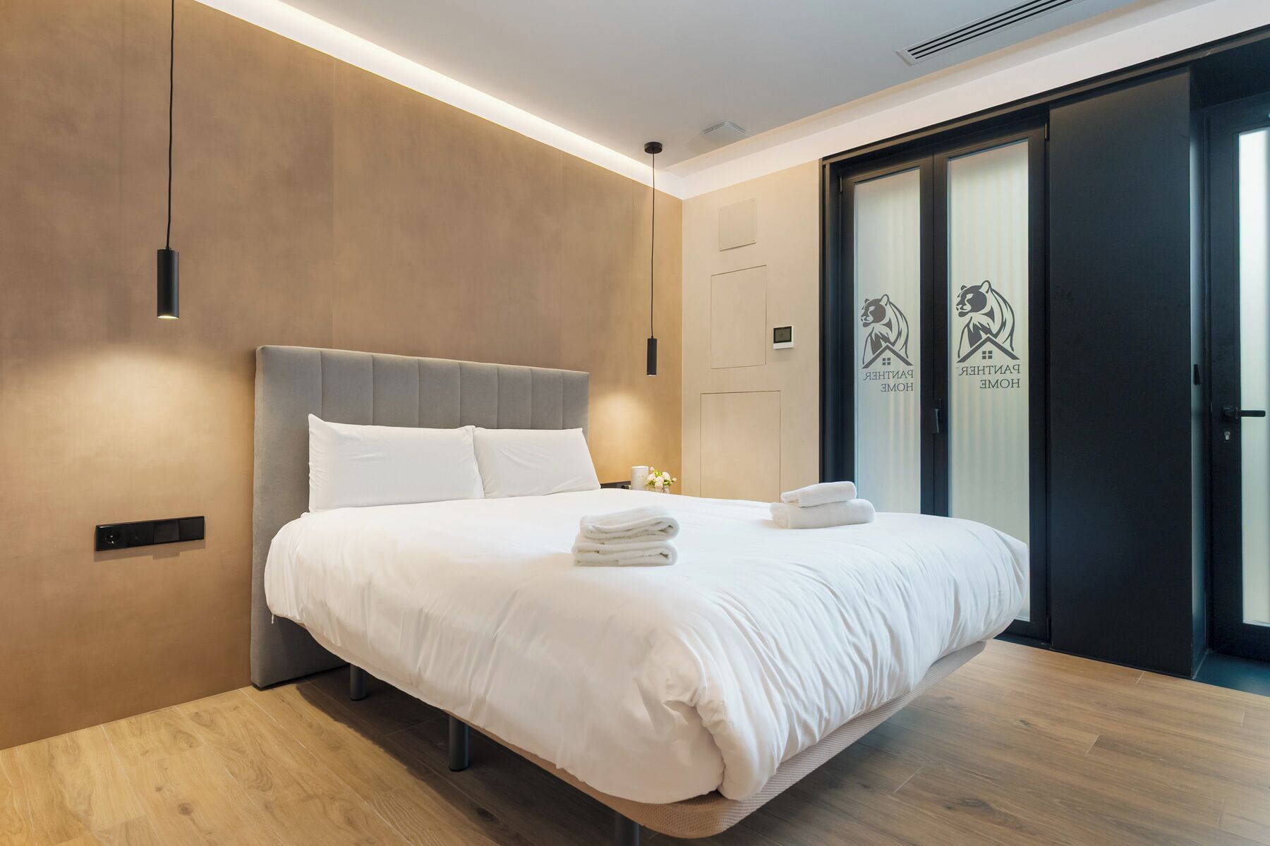 Une chambre d’hôtel moderne dotée d’un grand lit, d’une table de chevet, d’un panneau de commande fixé au mur et d’une porte coulissante ornée d’un logo.