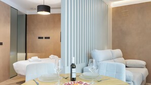 A modern hotel room with a bed, a seating area with a sofa and armchairs, a dining table with a plate of food and wine glasses, and a vertical light panel. - Panther Home Puerta Osario (Casco Antiguo)