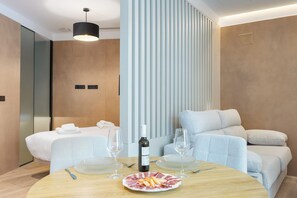 Una habitación de hotel moderna con una cama, un área de descanso con un sofá y sillones, una mesa de comedor con un plato de comida y copas de vino, y un panel de luz vertical.