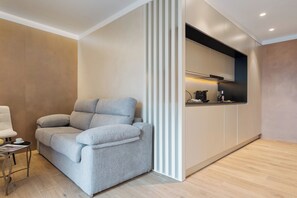 Una sala de estar moderna con un sofá gris, una mesita con una taza y una revista, y una zona de cocina con una cafetera.