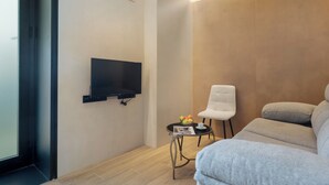 A modern living room with a flat-screen TV mounted on the wall, a white chair, a grey sofa, and a small round table with a fruit bowl and a magazine. - Panther Home Puerta Osario (Casco Antiguo)