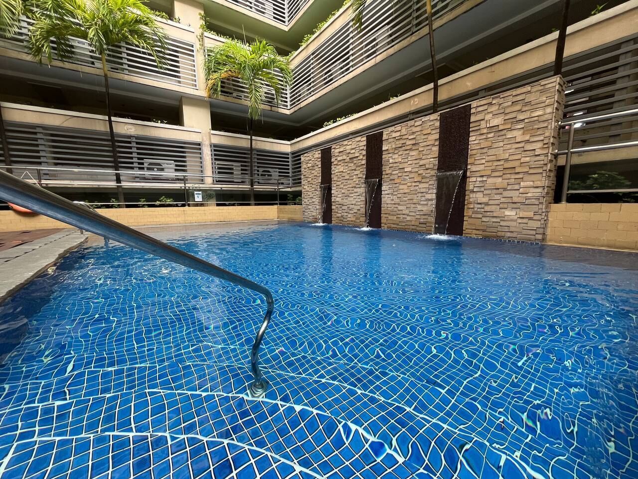 An indoor swimming pool with a blue mosaic floor, surrounded by a modern building with balconies and a stone wall.