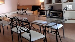 Zona pranzo con tavolo in vetro e sedie in metallo, soffitto in legno e parete bianca con mensole.