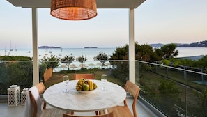 A balcony with a round table, chairs, a wicker light fixture, and a view of the sea with boats. - Ammos luxury apartments (Skiathos)