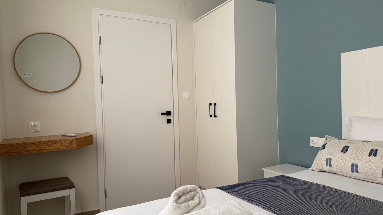 Een slaapkamer met een bed, nachtkastje, spiegel, kledingkast en een bankje.