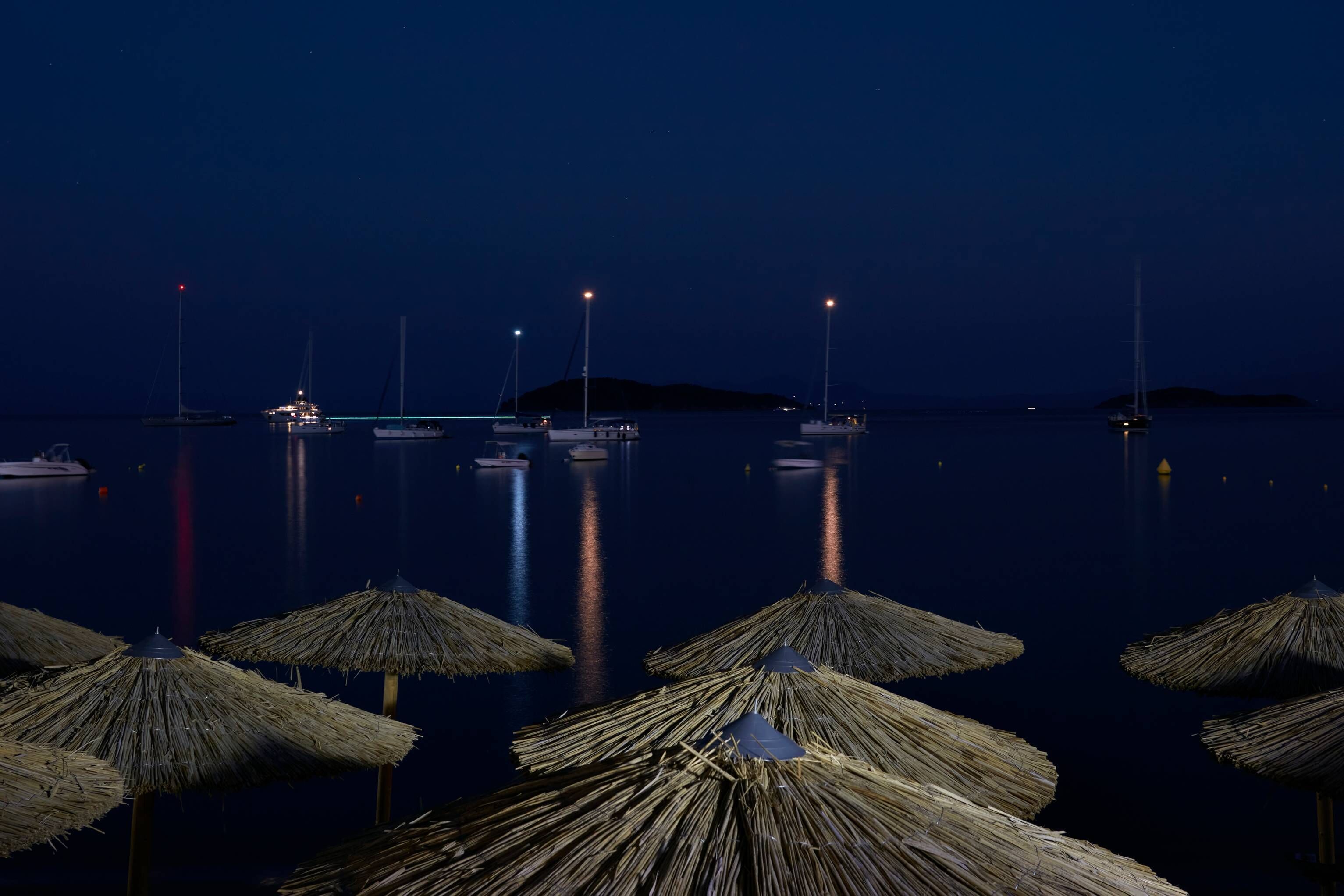 Una marina nocturna con barcos anclados en aguas tranquilas, iluminada por luces lejanas y sombrillas de paja en primer plano.