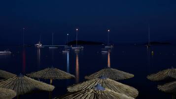 Una marina nocturna con barcos anclados en aguas tranquilas, iluminada por luces lejanas y sombrillas de paja en primer plano.