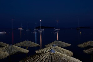 A nighttime marina with boats anchored in calm waters, illuminated by distant lights, and straw umbrellas in the foreground.