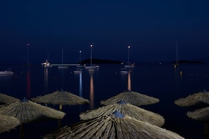 Una marina nocturna con barcos anclados en aguas tranquilas, iluminada por luces lejanas y sombrillas de paja en primer plano.