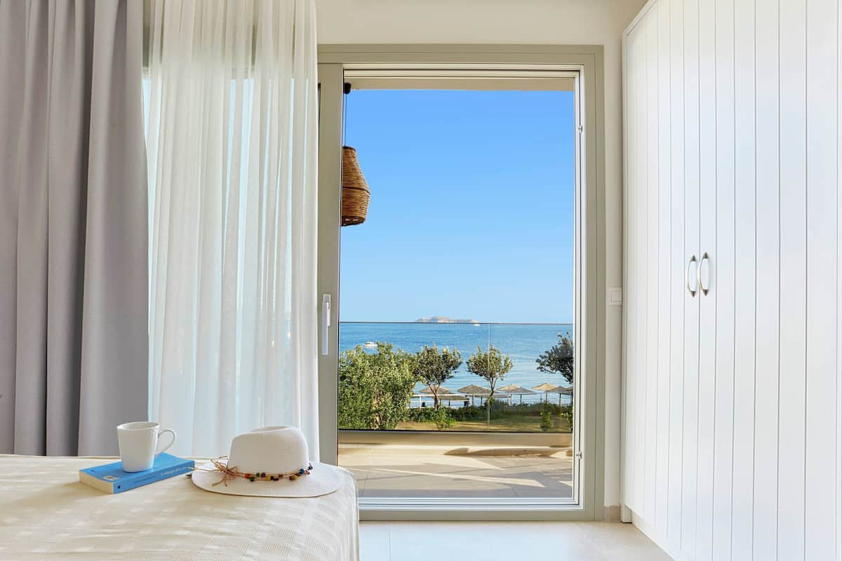A room with a bed, a white hat, a cup, and a book on a table. There is a view of the sea and a beach through a glass door.