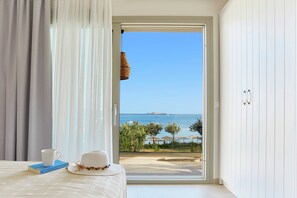 A room with a bed, a white hat, a cup, and a book on a table. There is a view of the sea and a beach through a glass door. - Ammos luxury apartments (Skiathos)