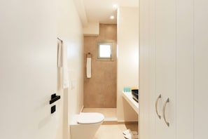 A narrow bathroom with a toilet, a towel rack, a small window, and a shower area. - Ammos luxury apartments (Skiathos)