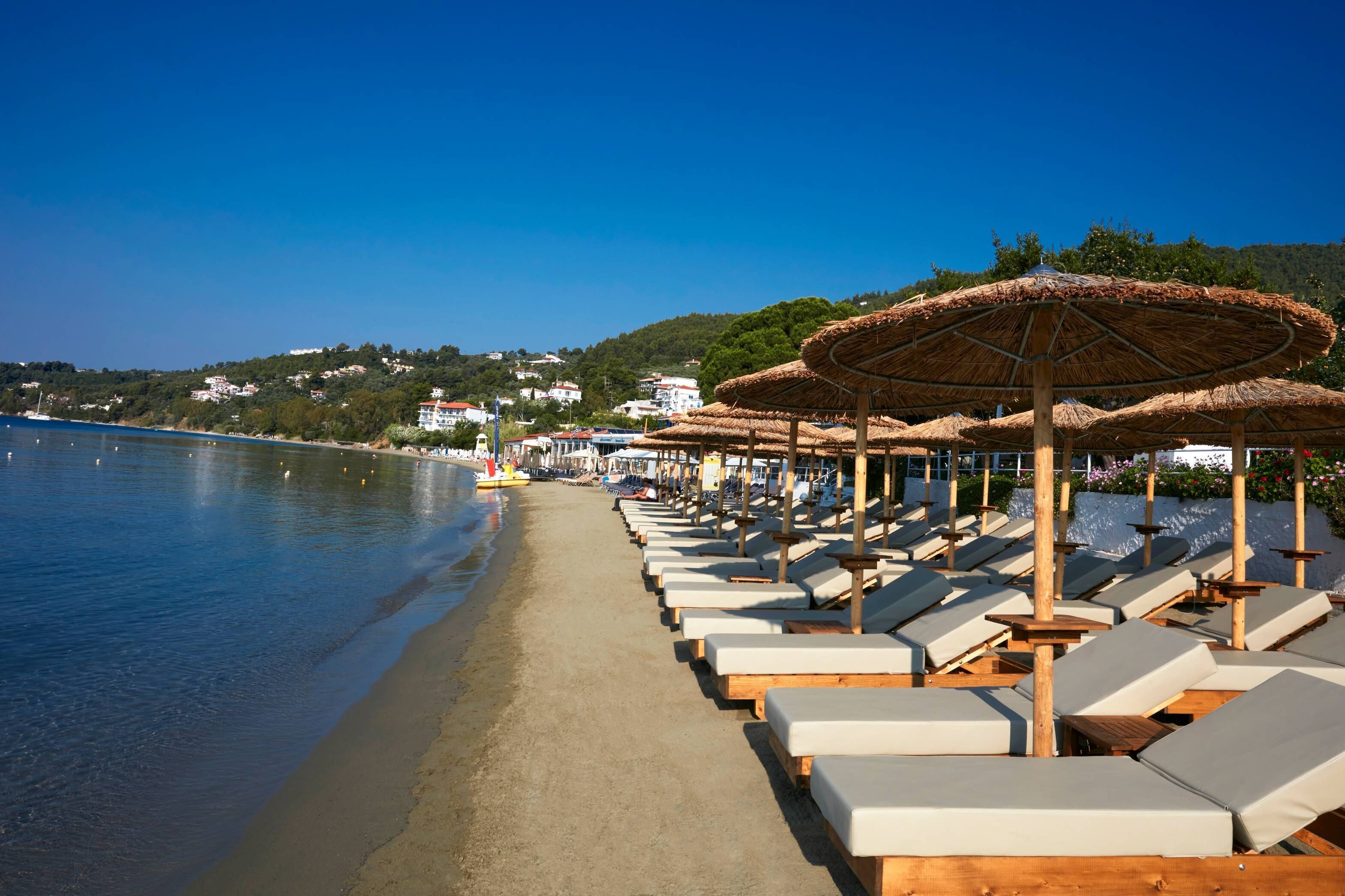 A beach with rows of sun loungers and thatched umbrellas, a clear sky, and a calm sea.