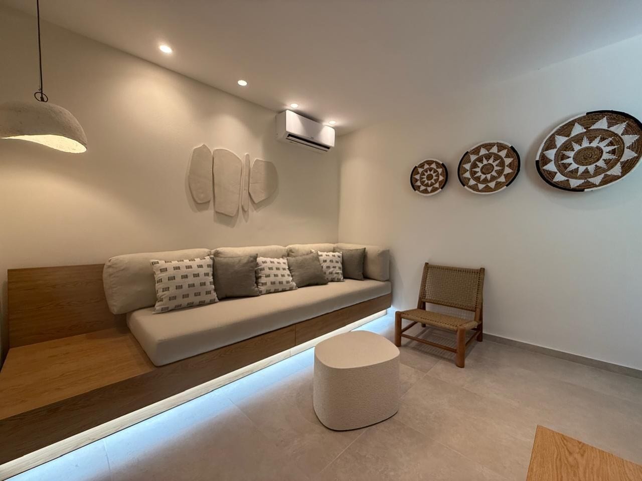 A modern living room with a sofa, a round ottoman, a wooden chair, and wall-mounted decor.