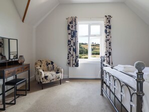 A bedroom with a bed, a chair, a dresser, a mirror, and a window with curtains. - Pwll Glas (Caersws)