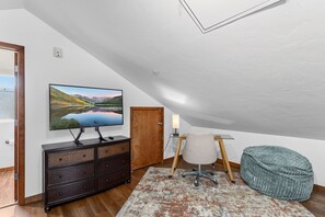 Ein Zimmer mit Fernseher auf einer Kommode, einem Schreibtisch mit Lampe, einem Sessel und einem Sitzsack.