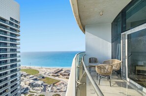 Un balcon avec deux fauteuils en osier, une balustrade en verre et une vue sur l’océan et une plage.