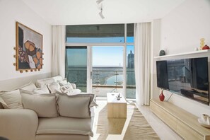 A modern living room with a sofa, a coffee table, and a television. There is a large window with a view of the city.