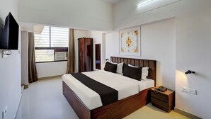 A hotel room with a bed, bedside table, a flat-screen TV mounted on the wall, a window with curtains, and a wall-mounted air conditioning unit. - Super OYO Collection O Prayag Junction (Prayagraj)
