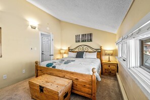 Ein Schlafzimmer mit einem hölzernen Bett, Nachttischlampen, einem Nachttisch, einer Holzkiste, einer Tür und einem Fenster mit Vorhängen.