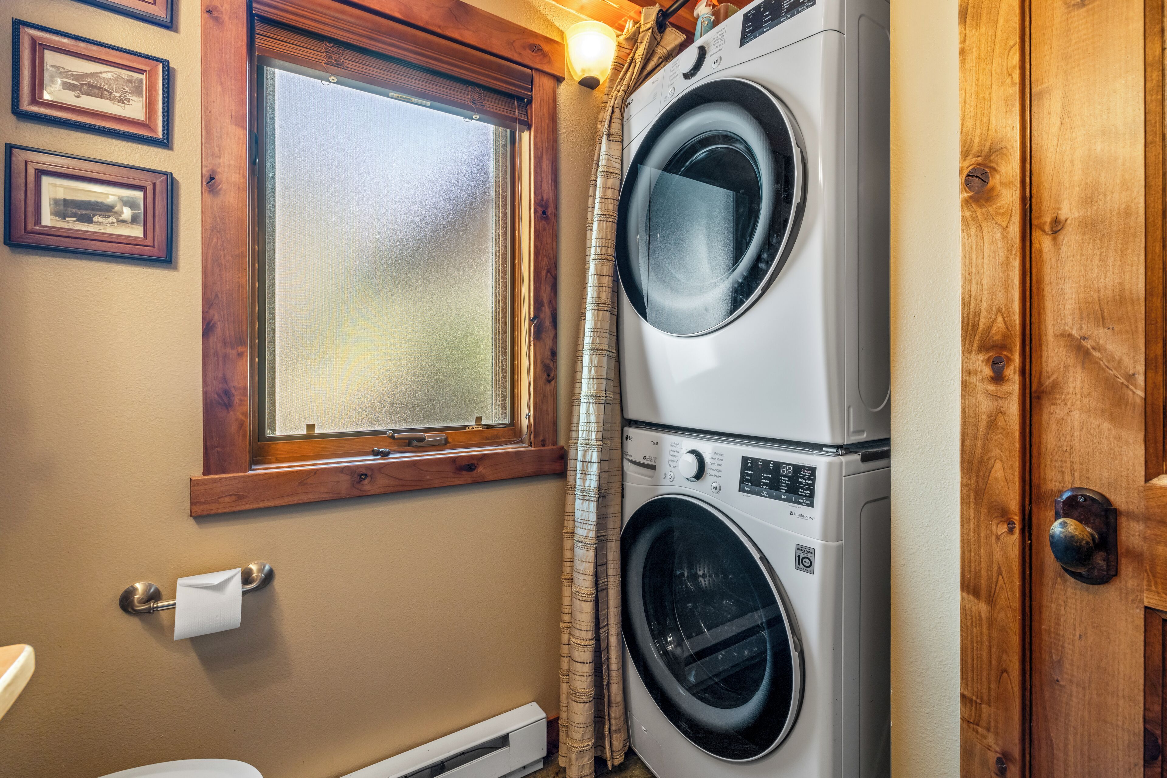 A laundry room with two stacked front-loading washing machines, a window with a wooden frame, and a door with a wooden finish.