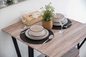 Ein Esszimmertisch ist gedeckt mit Tellern, Schüsseln, Besteck und Gläsern, dazu ein gewebter Korb und ein Blumentopf.