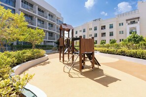 Ein Spielplatz mit Klettergerüsten und einer Rutsche, umgeben von einem begrünten Bereich mit Büschen und Bäumen, vor einem mehrstöckigen Wohngebäude.