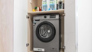 A compact Hisense washing machine in a small laundry room with a shelf above containing bottles of water and cleaning products. - StayShort - Contemporary Studio Sleeps 2 in JVC (JVC)