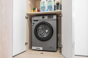 Eine kompakte Hisense Waschmaschine in einem kleinen Hauswirtschaftsraum mit oberhalb angebrachtem Regal, das Wasserflaschen und Reinigungsmittel enthält.