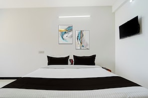 Habitación de hotel con una cama grande, dos almohadas blanco y negro, un televisor de pantalla plana montado en la pared y dos cuadros abstractos sobre la cama.