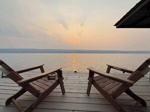 Eine Holzterrasse mit zwei Liegestühlen vor einem ruhigen Gewässer bei Sonnenuntergang.