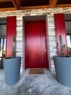A red door with a stone wall backdrop, two large potted cacti, and a doormat with 'hello' written on it.