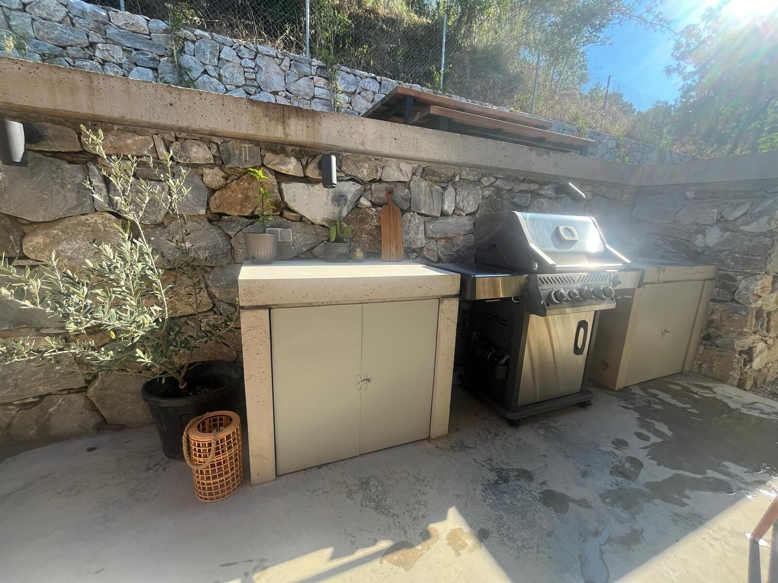 An outdoor kitchen with a stone wall backdrop, a built-in grill, and a trash can.