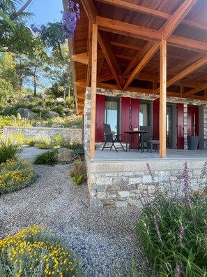 A covered outdoor seating area with stone walls and red shutters, surrounded by a gravel path and various plants. - Elmalı Guest House 
A simple, serene, and deeply peaceful life awaits you! (Muğla)