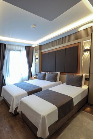 Habitación de hotel con dos camas, piso de madera, un ventanal grande con cortinas y un diseño moderno en el techo.