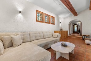 A spacious living area with a beige sectional sofa, a round coffee table, and a wooden cabinet. There are two windows with wooden frames and a ceiling with exposed beams. - Villa Cococinel by Sur la mer (Salernes)