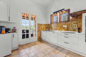 A kitchen with white cabinets, a tiled backsplash, and a window with a view of greenery. - Villa Cococinel by Sur la mer (Salernes)