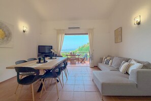 A living room with a sofa, a dining table with chairs, a TV, and a view through a window. - Sea View over Portisco Bay - Spacious 3BR Home - Clima (Porto Rotondo)