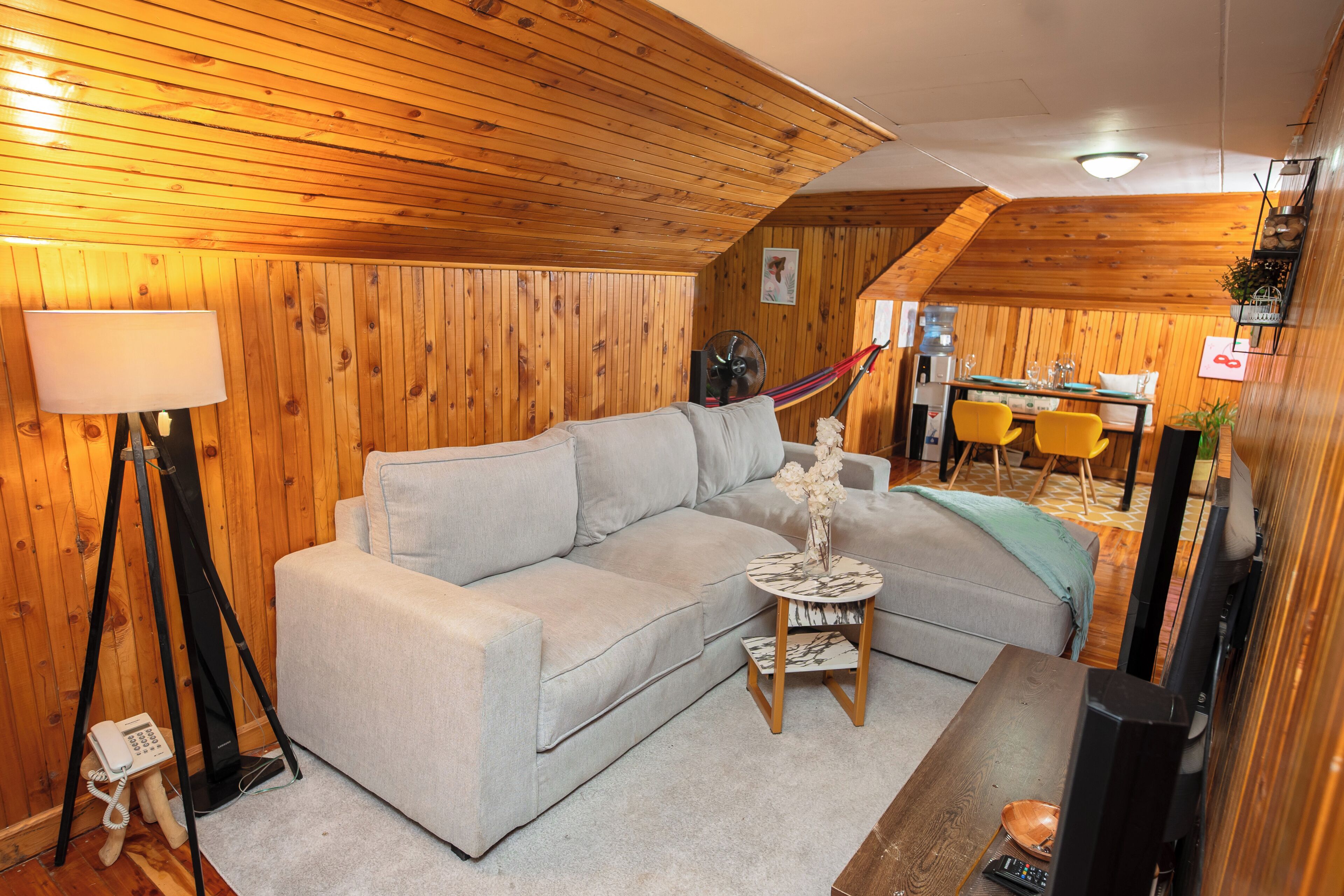 A cozy room with a wooden ceiling, a grey sofa, a small round table with a vase, and a dining area with yellow chairs.
