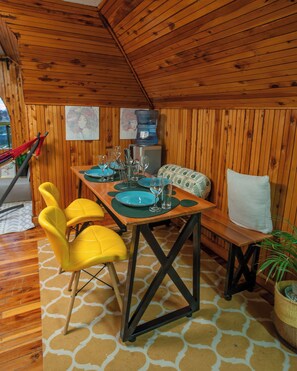 Interior de madera con una mesa de comedor preparada para cuatro, dos sillas amarillas, un banco con cojín blanco y un dispensador de agua.