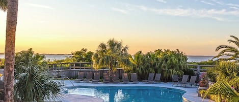 A pool area with lounge chairs and palm trees, overlooking a body of water.
