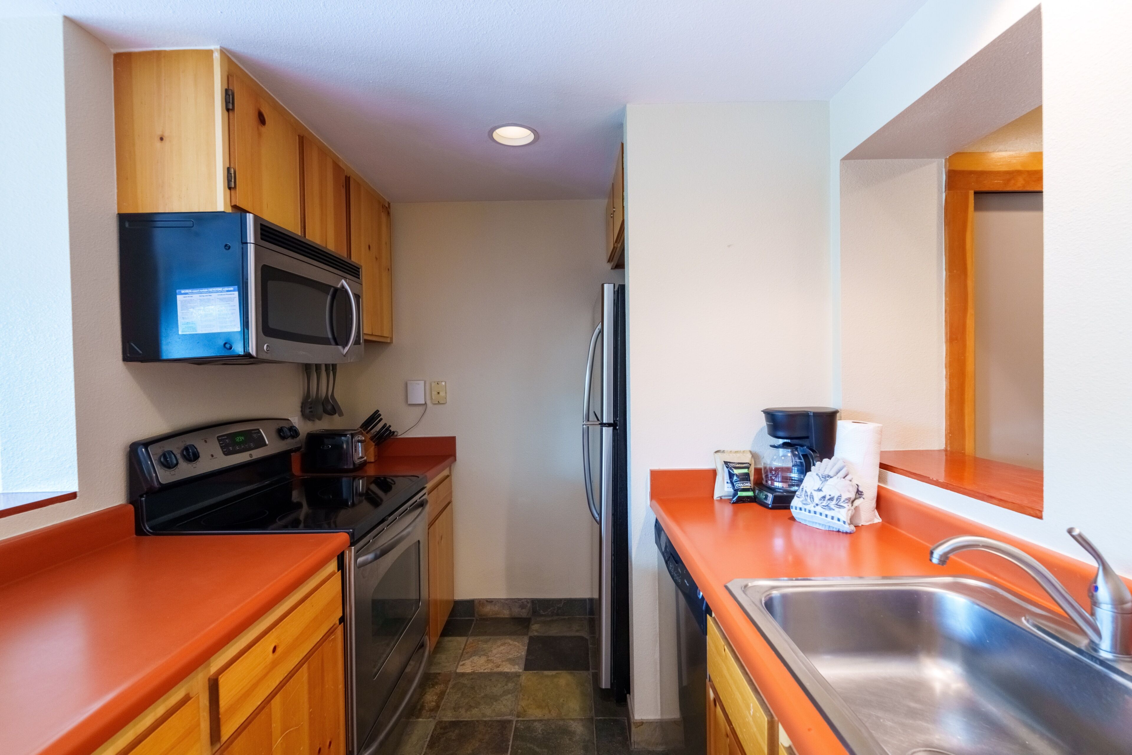 A compact kitchen with wooden cabinets, a stainless steel refrigerator, a microwave, a stove, and a sink.
