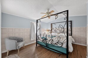 Un lit à baldaquin avec un ventilateur de plafond, un fauteuil gris et un plancher en bois dans une pièce aux murs bleu clair et aux boiseries blanches.