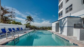 A swimming pool with blue water, surrounded by a white building with balconies, and a palm tree in the background. - Beach Villas 101 Beachfront Condo: Heated Pool, Walk To Dwtn 2 Bedroom Condo (Fort Myers Beach)