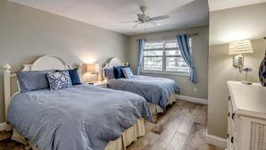 A bedroom with two beds, a ceiling fan, a window with curtains, and a bedside lamp. - Beach Villas 101 Beachfront Condo: Heated Pool, Walk To Dwtn 2 Bedroom Condo (Fort Myers Beach)