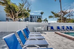 Zona de piscina con sillas de descanso y una mesa, rodeada por una valla blanca y árboles.